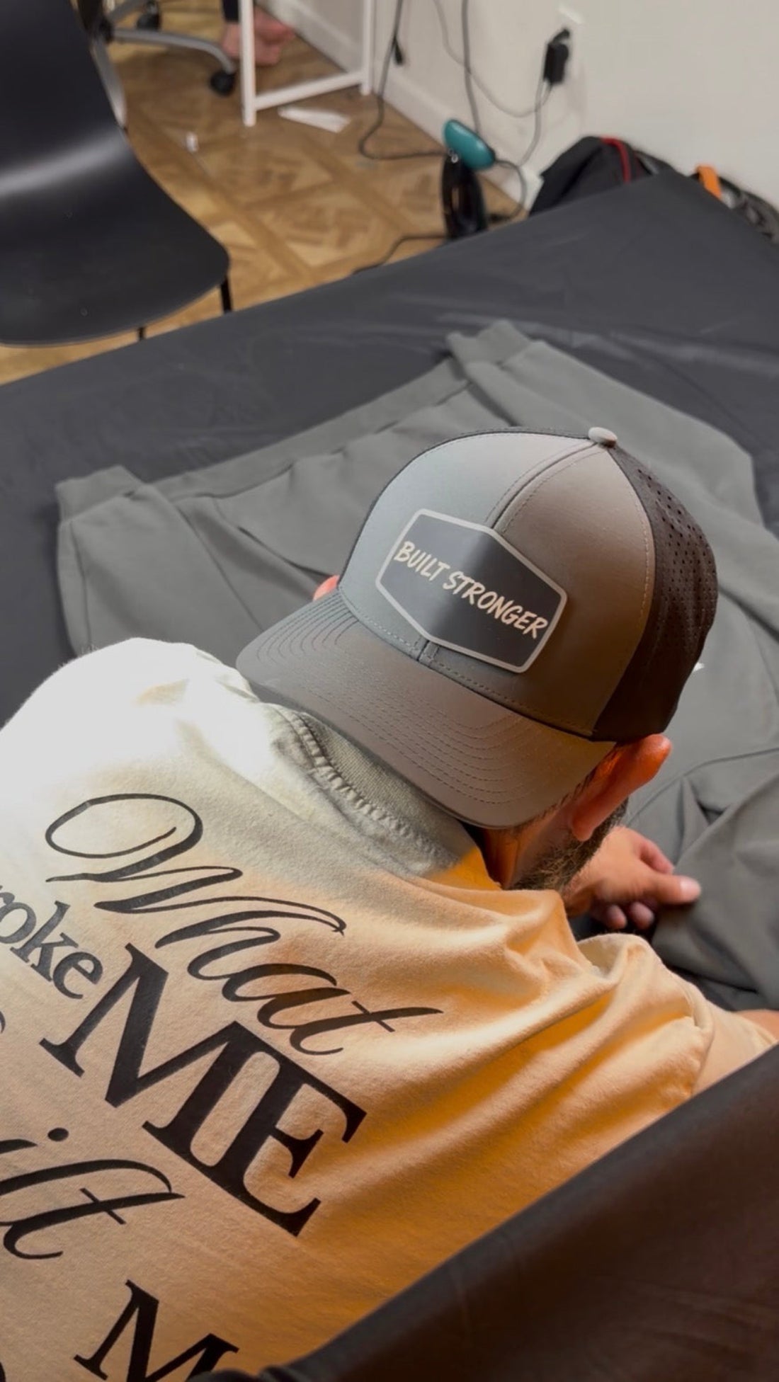 A man wearing a gray and black trucker hat with a 'Built Stronger' patch and a beige graphic t-shirt, seen from behind as he sits on a bed with a gray sweatshirt.