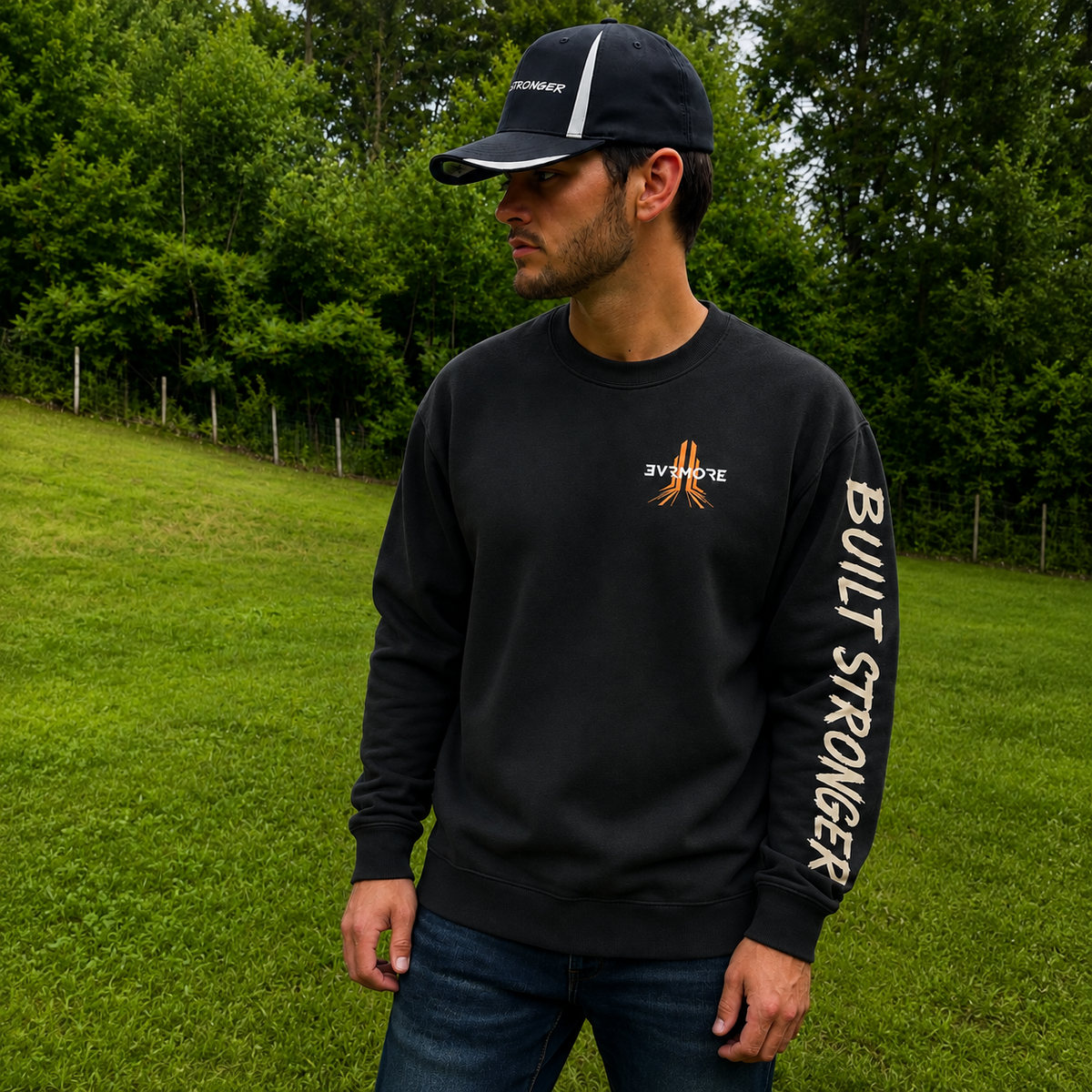 A man wearing a black crewneck sweatshirt with a small orange and white logo on the chest and 'BUILT STRONGER' printed vertically in white on the right sleeve, paired with a black baseball cap and blue jeans, standing on a grassy field with trees in the background.