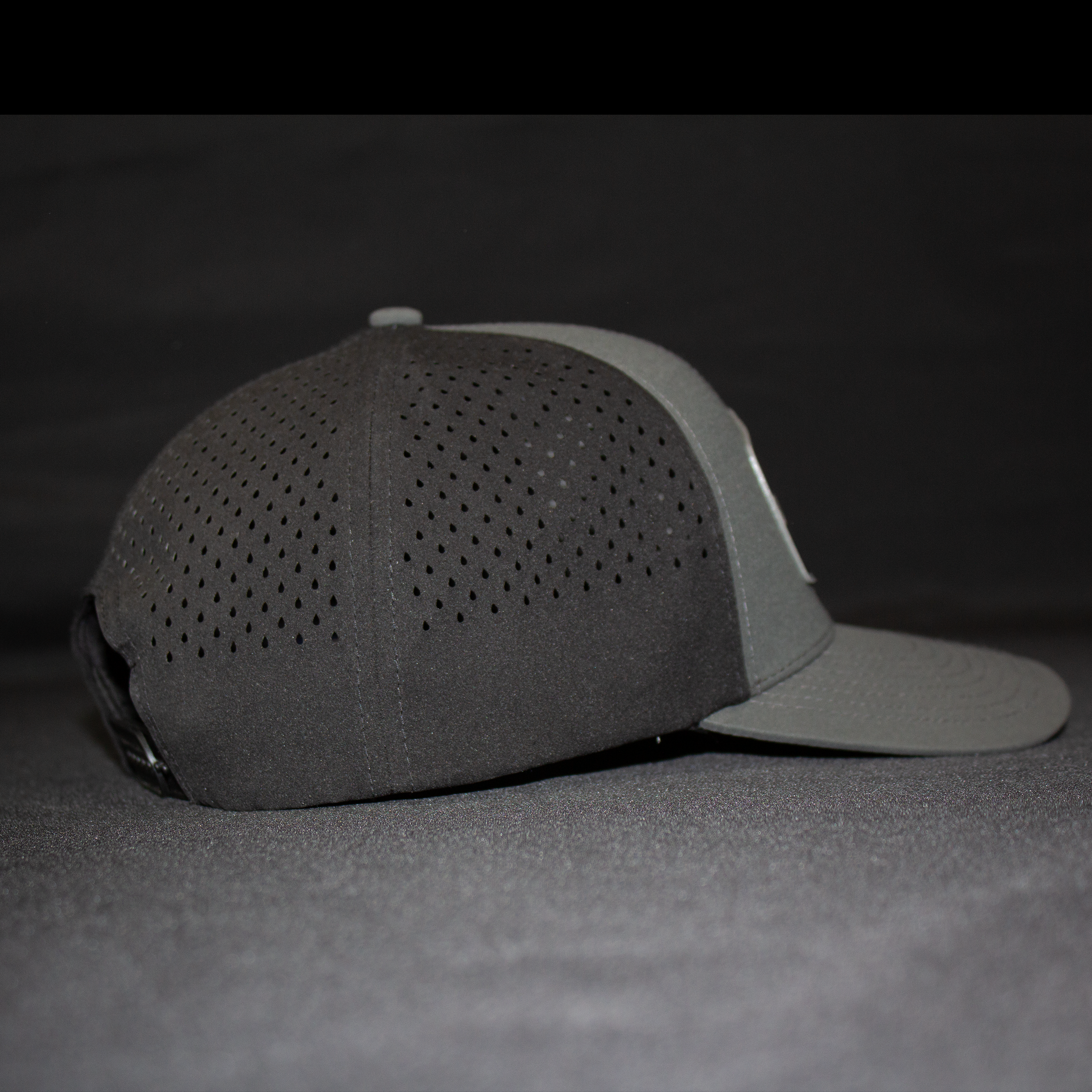 A dark gray and light gray baseball cap with perforated panels on the front, shown from a side profile against a dark gray background.