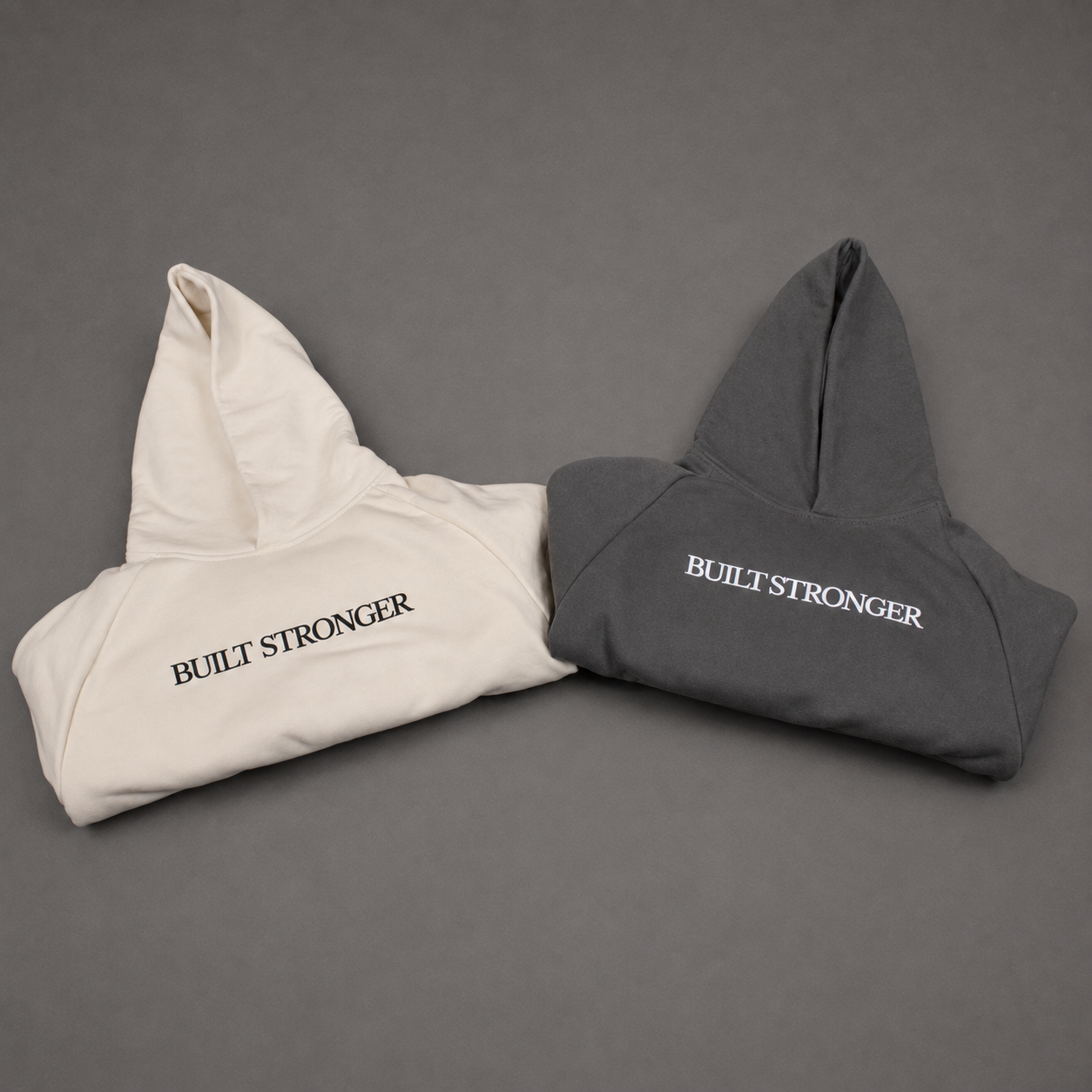 Two folded hoodies on a dark gray background, one in off-white with black 'BUILT STRONGER' text and one in dark gray with white 'BUILT STRONGER' text.