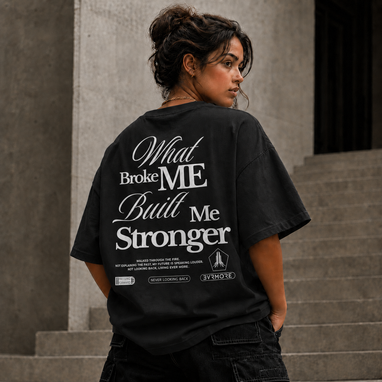 A woman wearing a black oversized graphic T-shirt with white text on the back reading 'What Broke ME Built Me Stronger' and smaller text below, photographed outdoors against concrete steps and a wall.