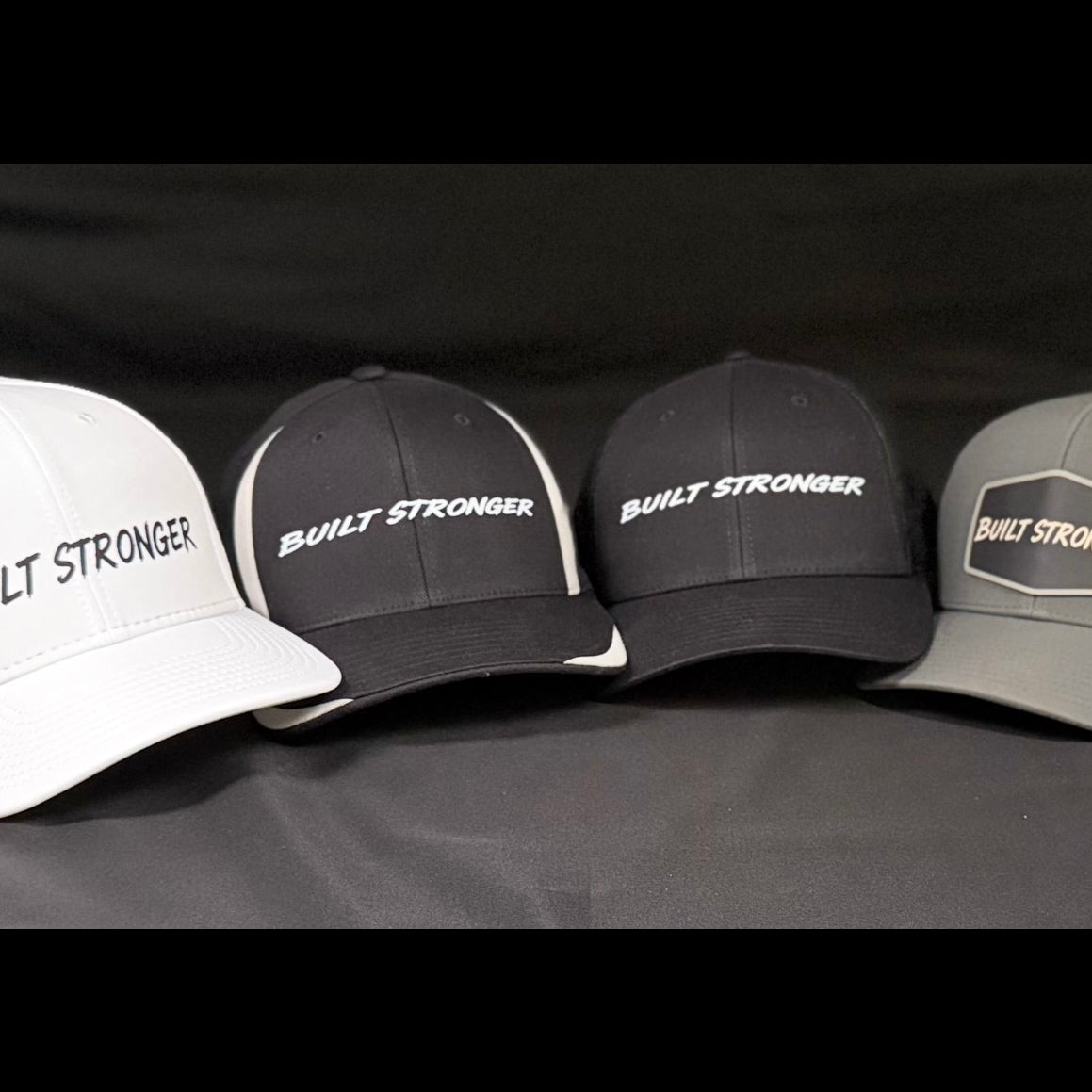 Four trucker-style baseball caps displayed side by side against a dark fabric backdrop. The caps are white, black, and gray, each featuring the text 'BUILT STRONGER' printed on the front panels.