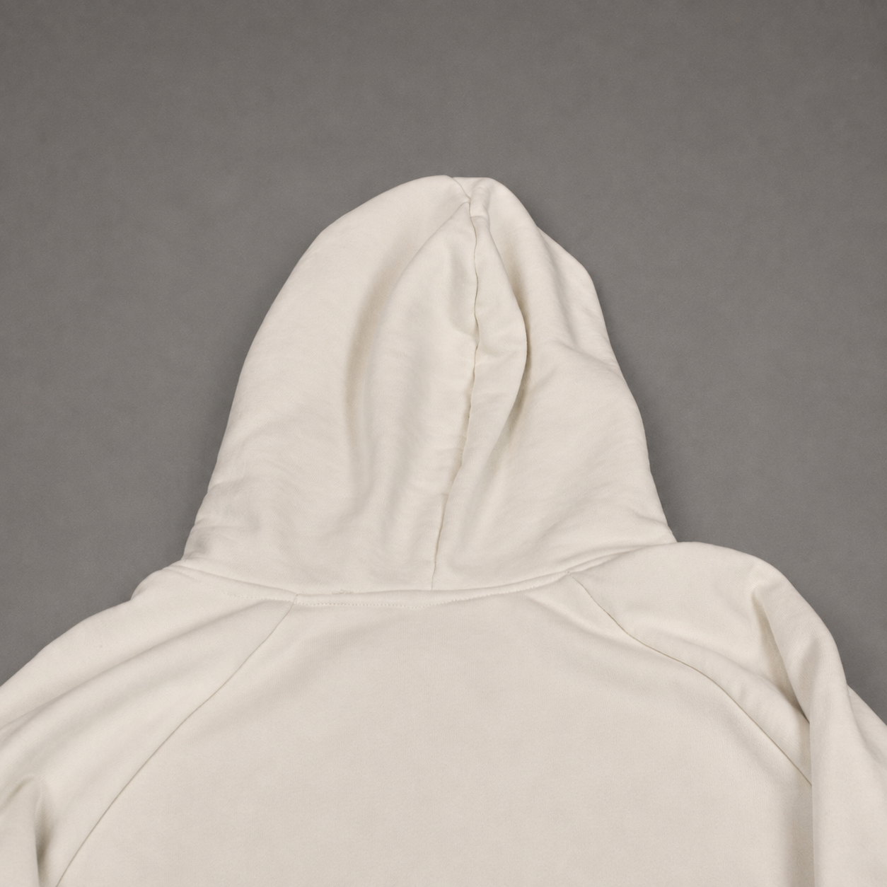 Close-up of the hood and upper back of a plain hoodie in a warm off-white or cream color, laid flat against a solid gray background.