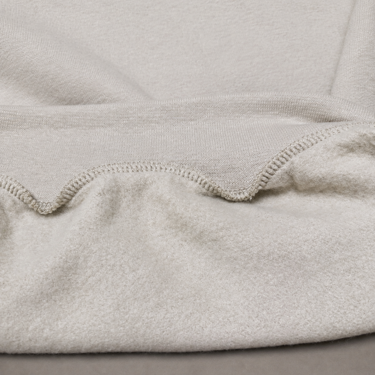Close-up of a soft knit fabric in a light warm gray or oatmeal tone, showing the texture and a folded hem with ribbed stitching.