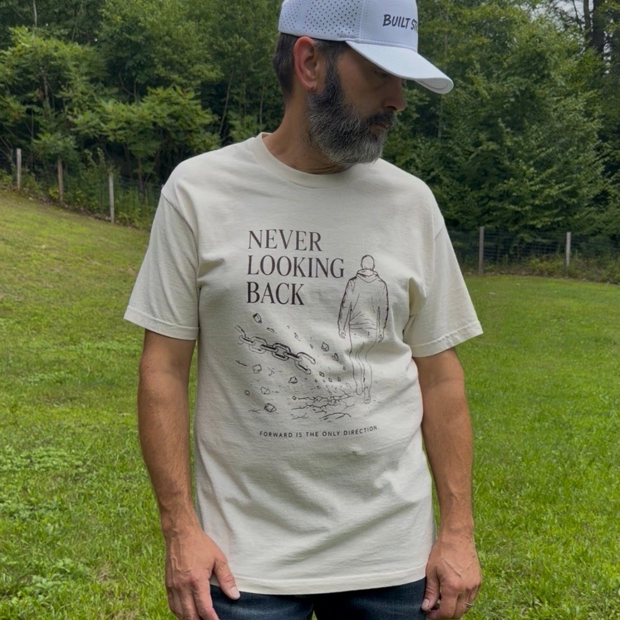 A man wearing a light beige graphic t-shirt with the text 'NEVER LOOKING BACK' and a line drawing, paired with blue jeans and a white cap, standing in a grassy outdoor setting.