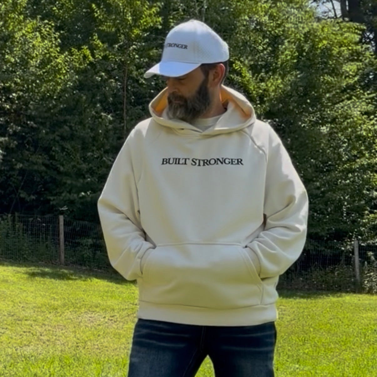 A man wearing a cream-colored hoodie with 'BUILT STRONGER' printed in black across the chest, dark blue jeans, and a white baseball cap, standing in a grassy outdoor park with trees in the background.