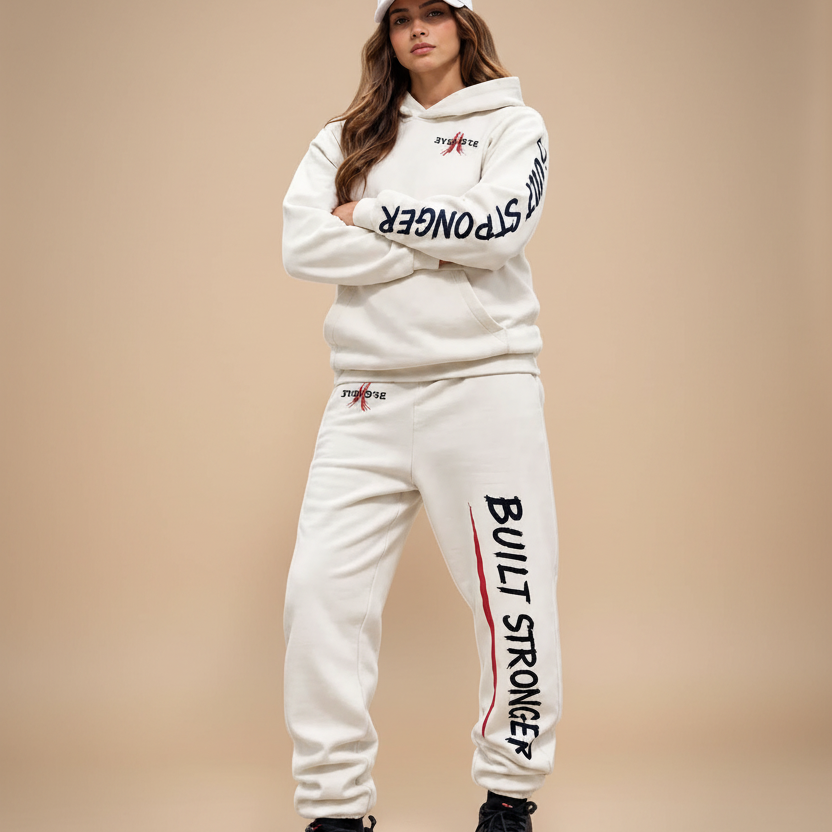 A woman modeling a matching off-white tracksuit with black and red 'Built Stronger' graphics, paired with a white cap and black sneakers with red accents, set against a solid black background.