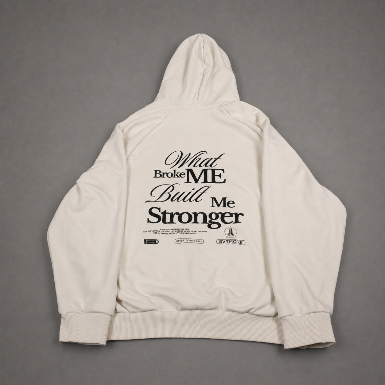 An off-white hooded sweatshirt shown from the back, featuring a black graphic text print that reads 'What Broke ME Built Me Stronger' with smaller text and logos underneath, laid flat on a gray background.