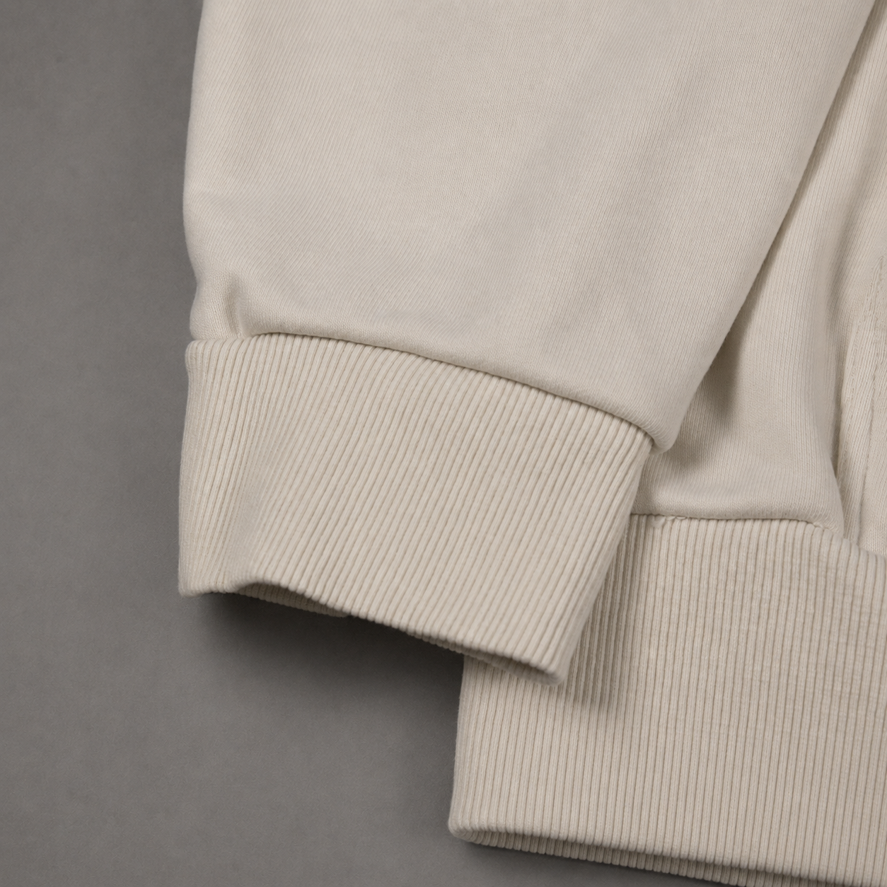 Close-up of a ribbed knit garment cuff in a light warm off-white or cream color, shown against a plain gray background.