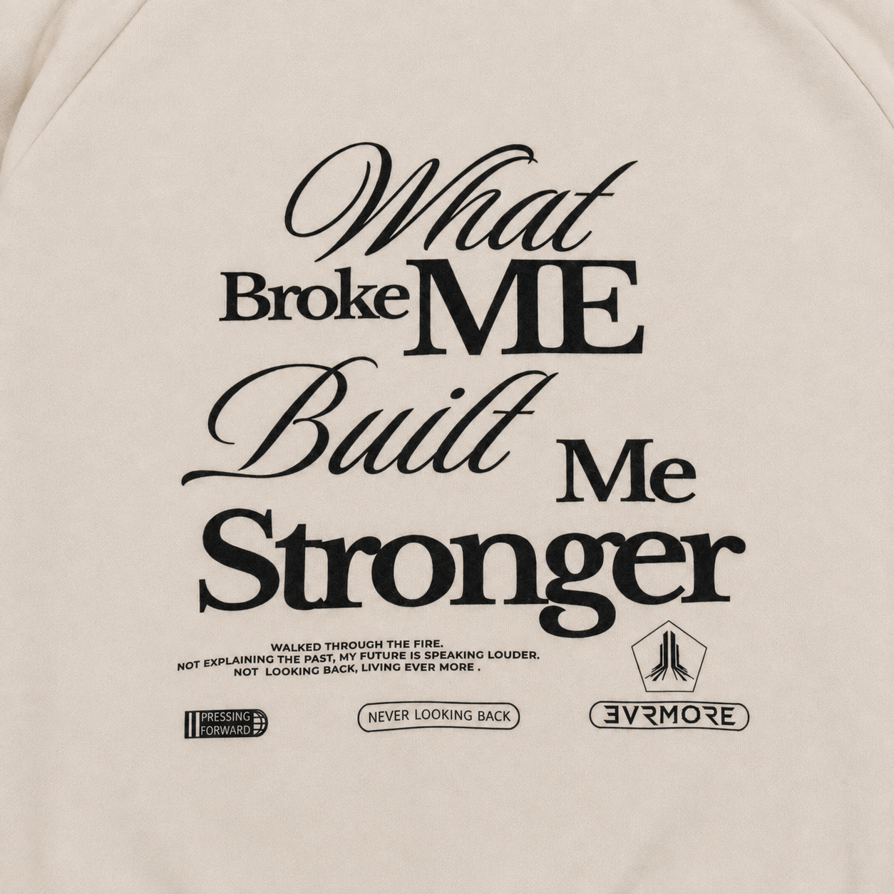 Close-up of a light beige or off-white garment with large black printed text reading 'What Broke ME Built Me Stronger' and smaller graphic text and logos below.