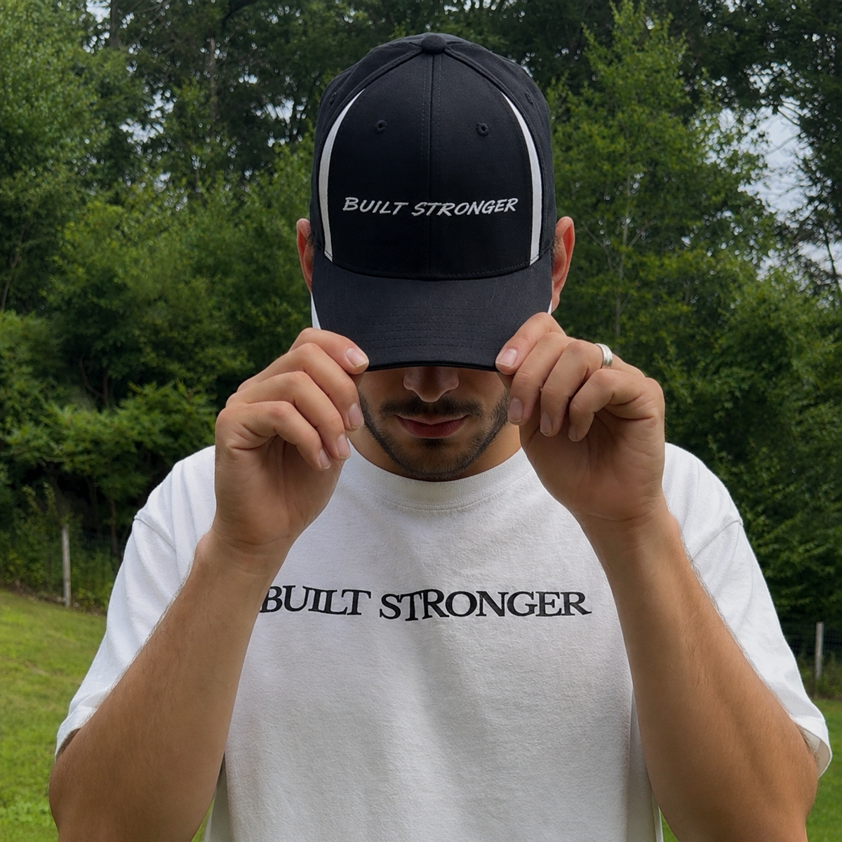 A man outdoors wearing a black baseball cap and a white t-shirt, both featuring the text 'BUILT STRONGER'. He is holding the brim of the cap down with both hands.