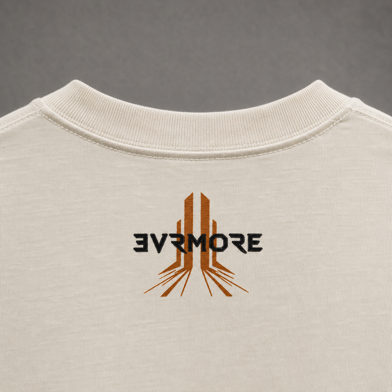 Close-up of the back of an off-white crewneck t-shirt featuring a stylized graphic with the text '3V7MORE' in black and a brown geometric emblem.