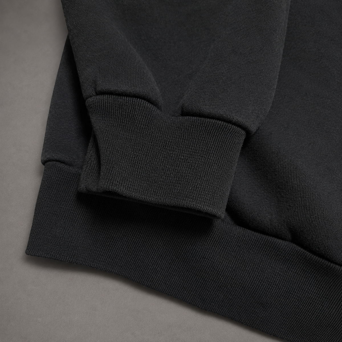Close-up detail of a dark charcoal gray ribbed knit garment, showing the sleeve cuff and hem against a neutral gray background.