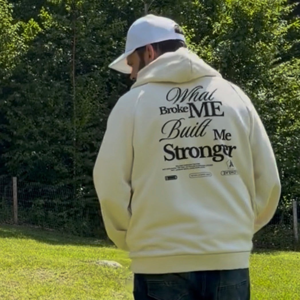 A man standing outdoors on a grassy field with trees in the background, wearing a cream-colored hoodie with black text on the back that reads 'What Broke ME Built Me Stronger', dark jeans, and a white baseball cap.