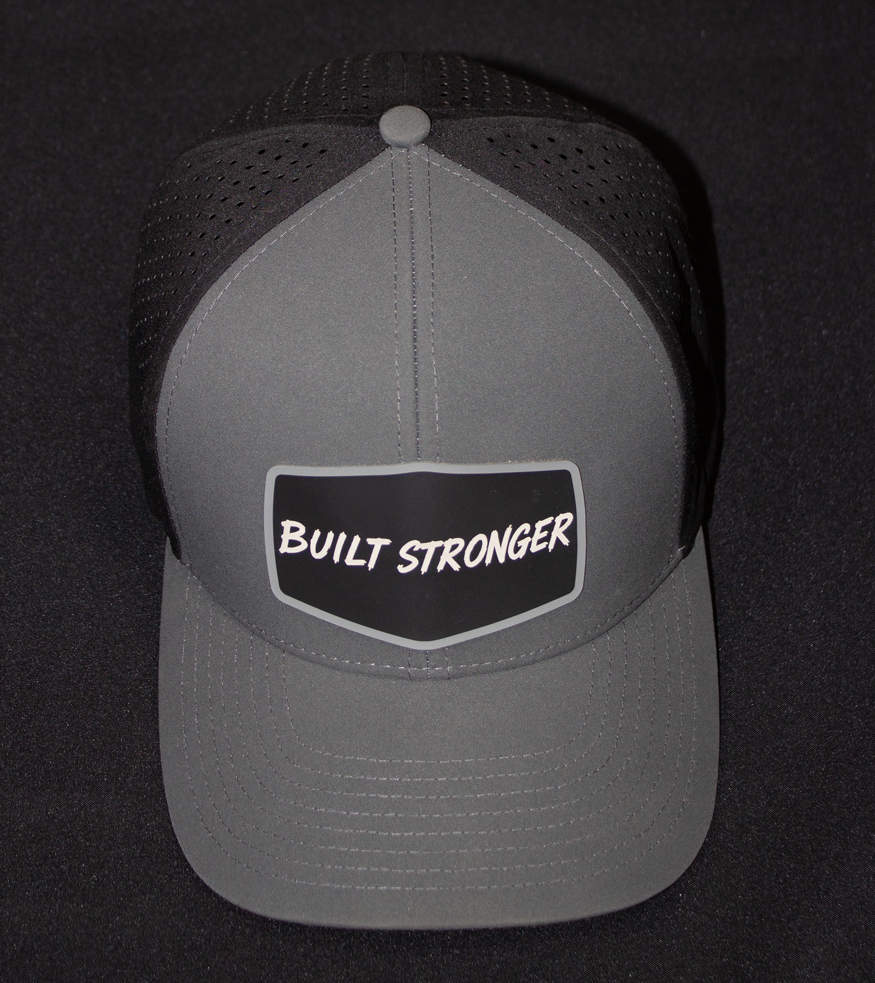 A gray and black trucker-style cap with a front patch reading 'BUILT STRONGER' in white text, photographed against a dark background.