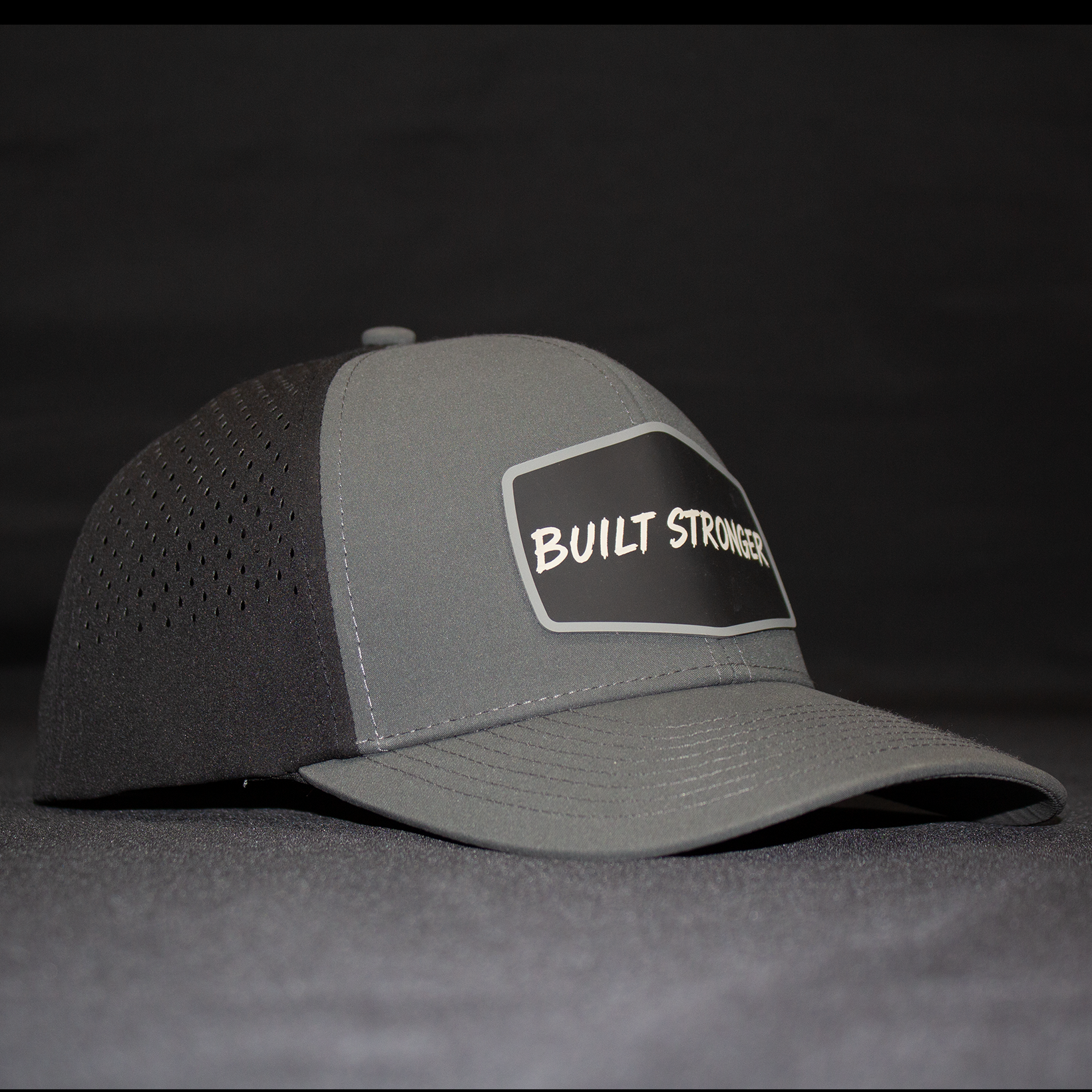 A gray trucker-style cap with perforated back panels and a black front patch reading 'BUILT STRONGER' in white, photographed against a dark gray backdrop.