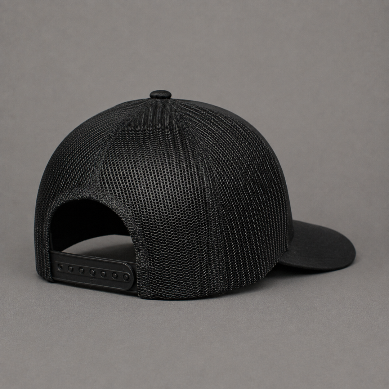 A black trucker-style baseball cap with a structured mesh back and curved brim, photographed from the rear on a neutral gray background.