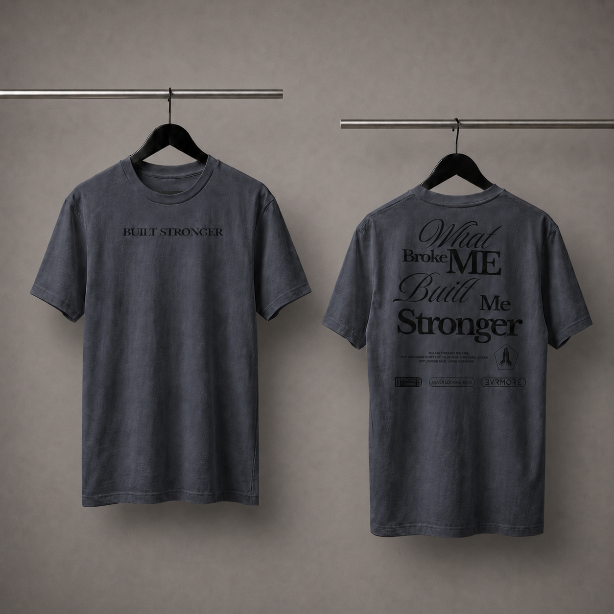 Two views of a dark gray washed t-shirt hanging on black hangers against a textured gray wall. The front view shows the text 'HURT STRONGER' in black on the chest, and the back view shows a large black graphic reading 'What Broke ME Built Me Stronger' with smaller text below.