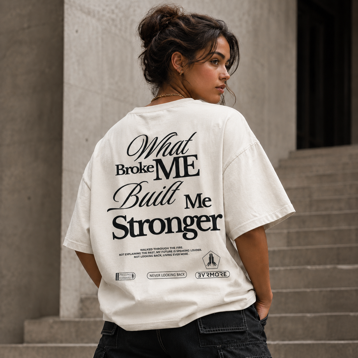 A woman wearing an oversized off-white graphic T-shirt with black text reading 'What Broke ME Built Me Stronger' and smaller text below, standing in front of concrete stairs.