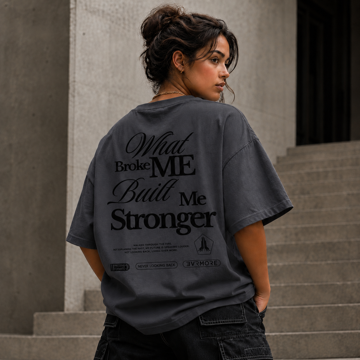 A woman wearing a dark gray oversized t-shirt with a black graphic text print on the back, standing on concrete stairs.
