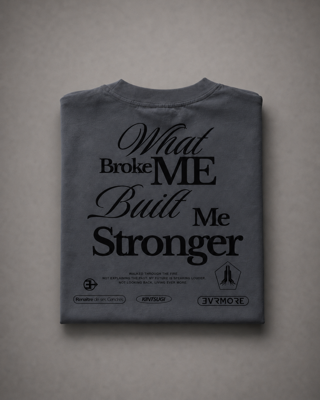 A folded dark gray t-shirt with black graphic text reading 'What Broke ME Built Me Stronger' and small printed logos/wordmarks at the bottom, displayed against a neutral gray background.