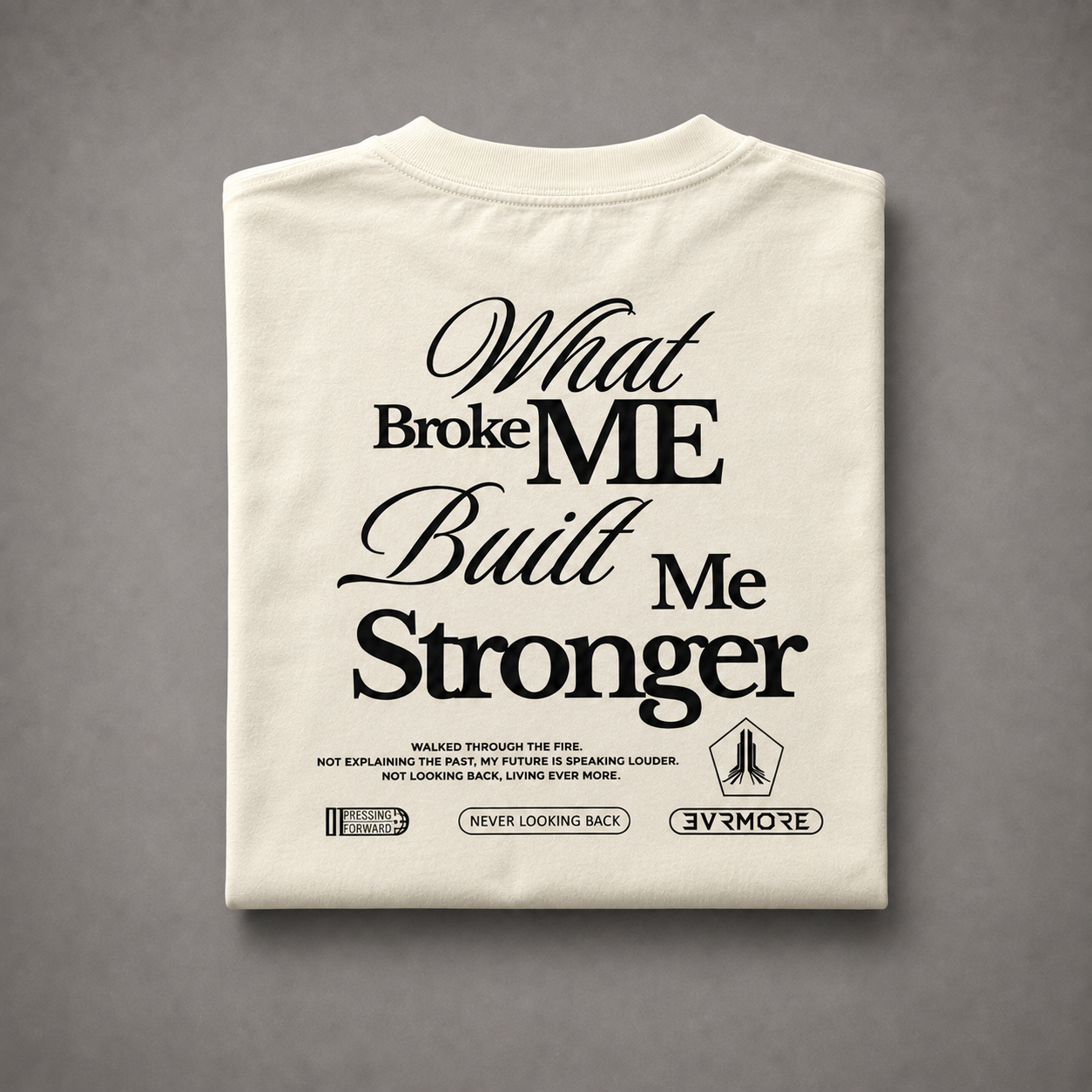 A folded off-white t-shirt with a black graphic text print reading 'What Broke ME Built Me Stronger' and smaller text below, displayed on a neutral gray background.