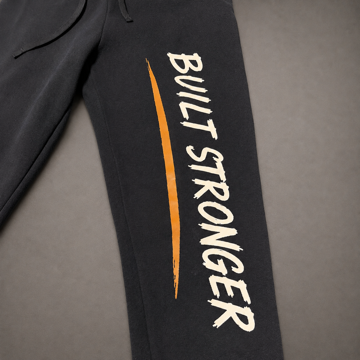 Close-up of black sweatpants laid flat on a gray background, featuring the words 'BUILT STRONGER' in white brush-style lettering and a vertical orange accent stripe on the leg.