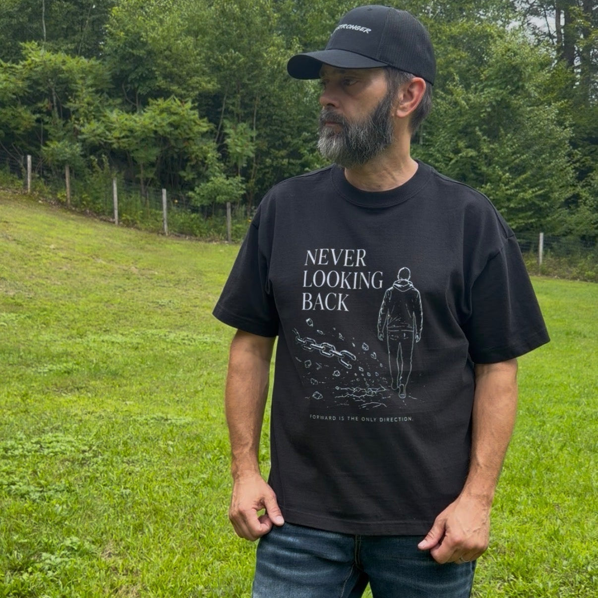 A man wearing a black graphic t-shirt with the text 'NEVER LOOKING BACK' and a silhouette, paired with dark blue jeans and a black baseball cap, standing in a grassy outdoor field.