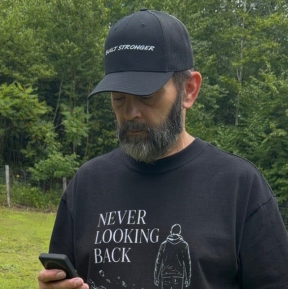 A man standing in a grassy field wearing a black t-shirt with a white graphic print that reads 'NEVER LOOKING BACK' and a black baseball cap, looking down at a phone.