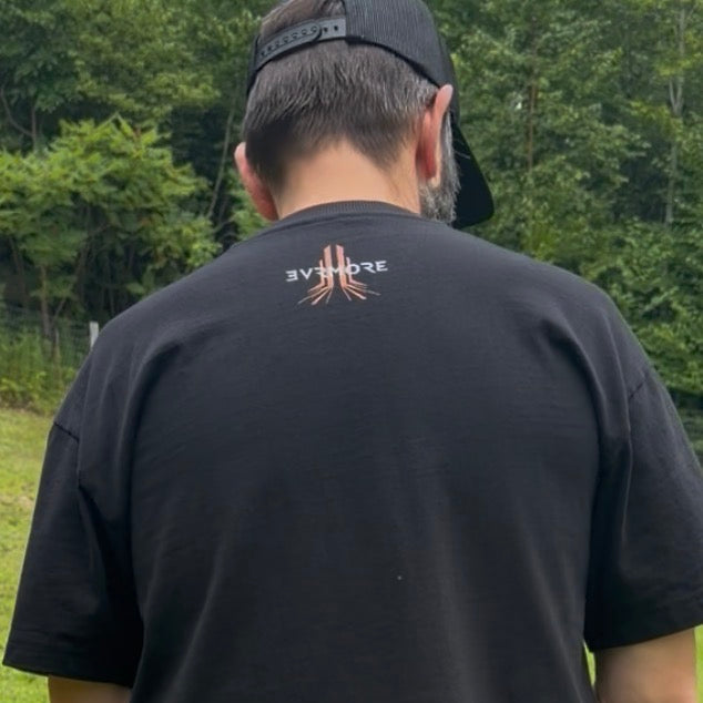 A man viewed from behind wearing a black t-shirt and a black baseball cap, standing outdoors in front of a green forest background. The back of the t-shirt features a small graphic logo with the word 'RESOLVE' in white and a red-orange campfire design.
