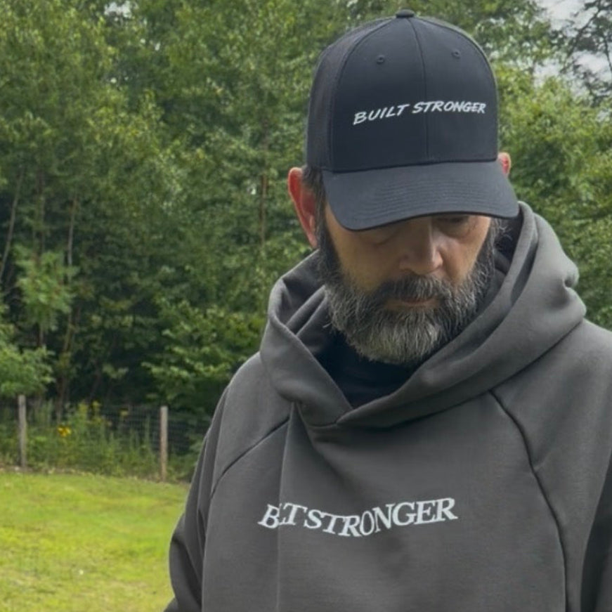 A man wearing a dark gray hoodie with 'BUILT STRONGER' printed in white on the chest, paired with a matching black baseball cap that reads 'BUILT STRONGER' and blue jeans, standing outdoors in a grassy field with trees in the background.