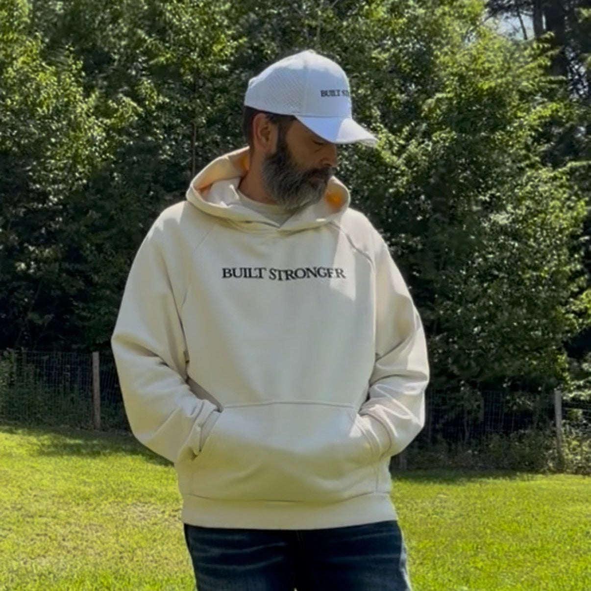 A man standing in a grassy outdoor park wearing a cream-colored hoodie with 'BUILT STRONGER' printed on the chest, dark jeans, and a white baseball cap.