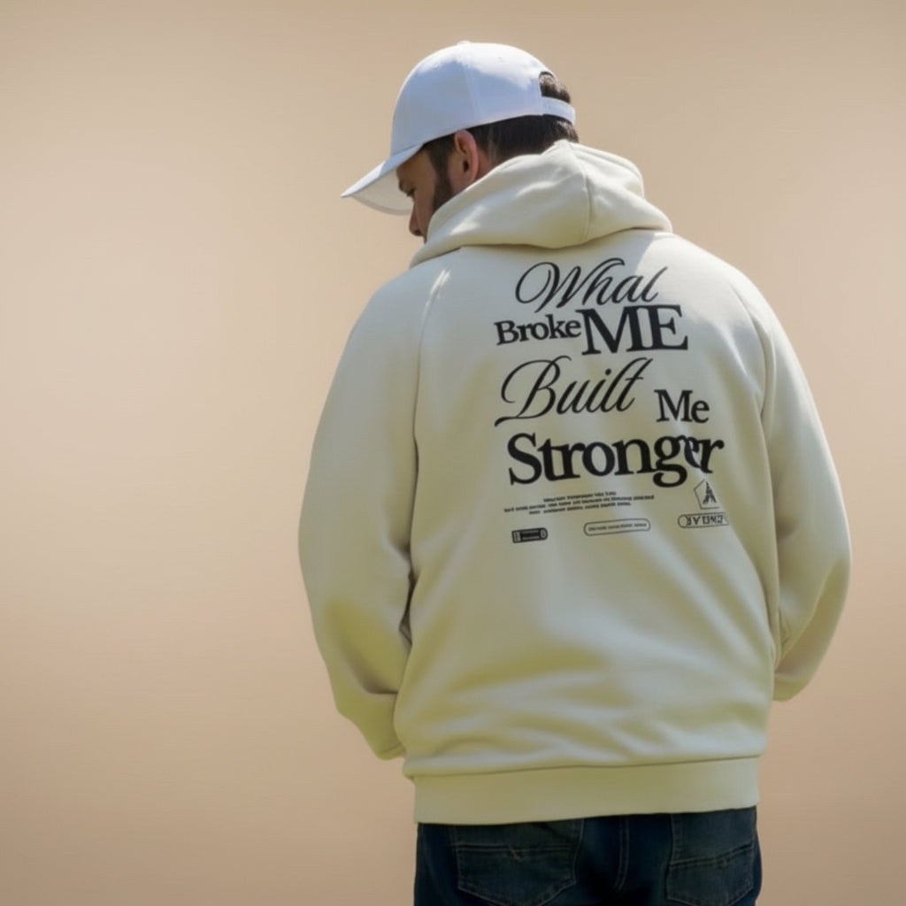 A man standing outdoors on a grassy field with trees in the background, wearing a cream-colored hoodie with black text on the back that reads 'What Broke ME Built Me Stronger', dark jeans, and a white baseball cap.