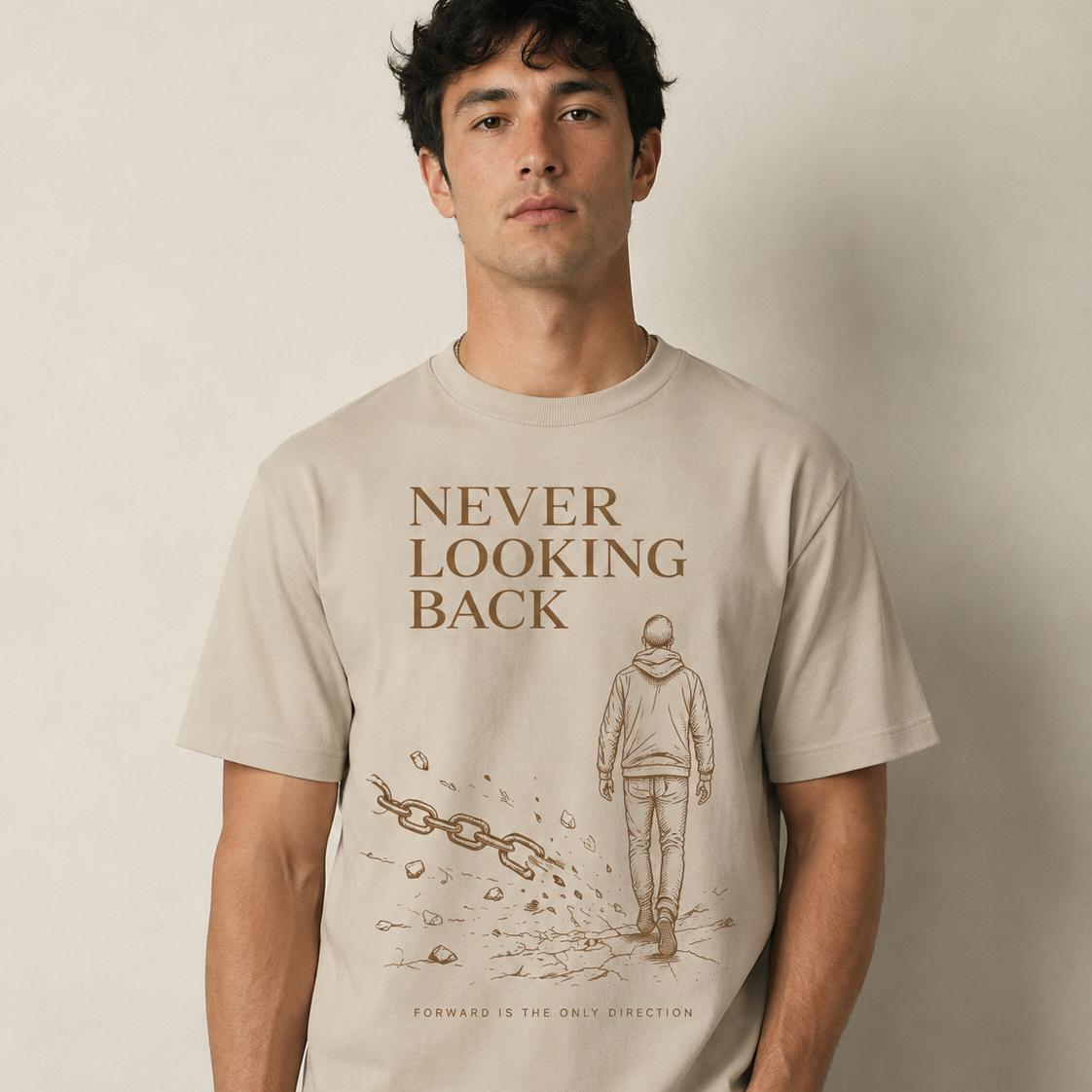 A man wearing a light beige graphic t-shirt with brown text reading 'NEVER LOOKING BACK' and an illustration of a person walking forward, paired with dark gray jeans, against a neutral beige background.