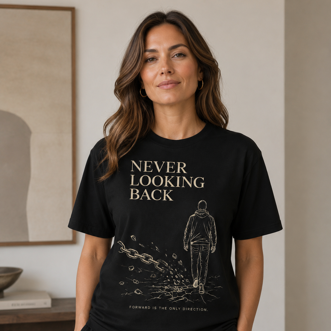 A woman wearing a black graphic t-shirt with a gold-toned print that reads 'NEVER LOOKING BACK' and 'FORWARD IS THE ONLY DIRECTION', paired with black sweatpants in a modern indoor setting.