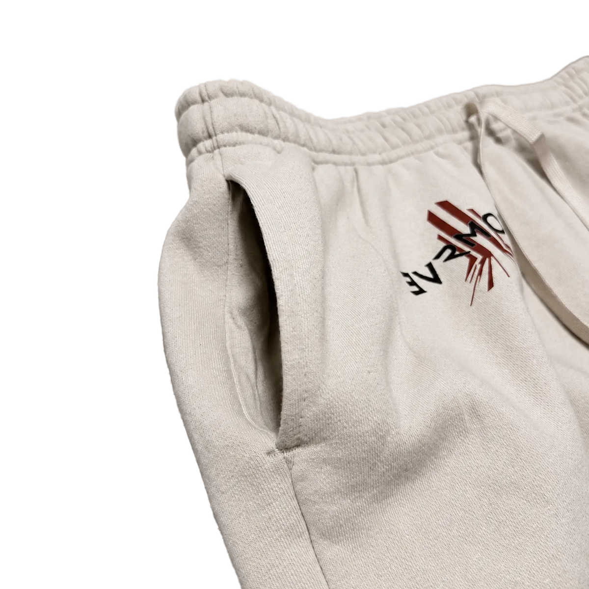 Close-up of off-white sweatpants with a drawstring waist and side pocket, featuring a black and red embroidered logo on the thigh, laid flat on a neutral gray background.
