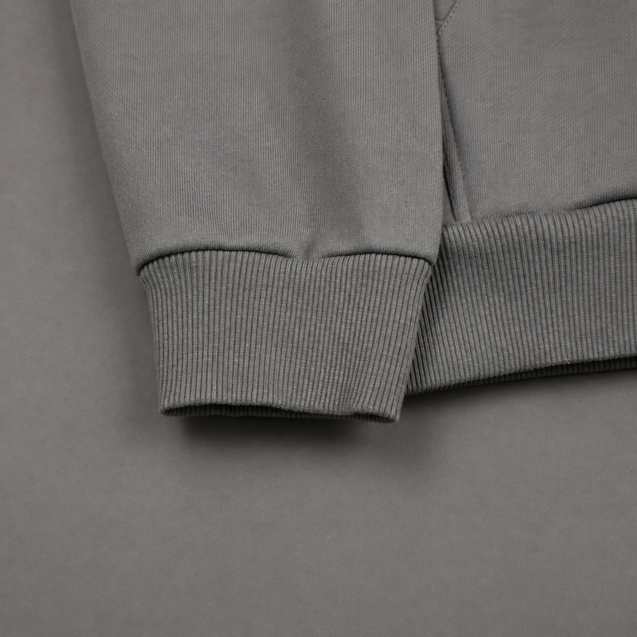 Close-up detail of a gray ribbed knit garment cuff, showing the fabric texture and folded sleeve construction on a matching gray background.