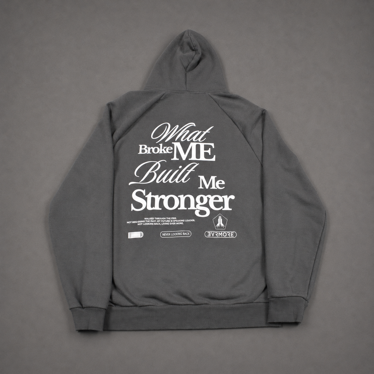A dark gray hoodie shown from the back, featuring a large white graphic text print that reads 'What Broke ME Built Me Stronger' with smaller text and logos underneath, laid flat on a gray background.