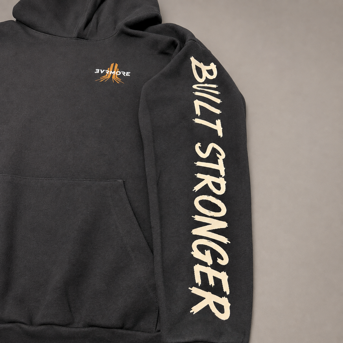 A close-up of a dark gray hoodie laid flat on a neutral gray background, featuring 'BUILD STRONGER' printed vertically in cream on the right sleeve and a small orange and white chest graphic.