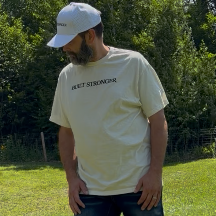 A man standing in a grassy outdoor park wearing a cream-colored t-shirt with black text that reads 'JUST STRONGER', dark blue jeans, and a white baseball cap.