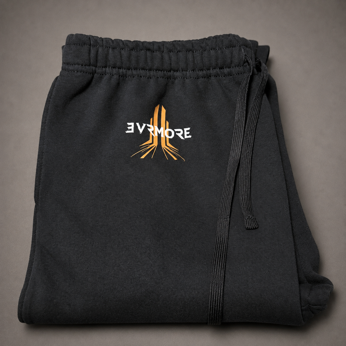 Black sweatpants folded flat on a neutral gray background, featuring a white and orange graphic logo on the front with the text 'EVNORE'.