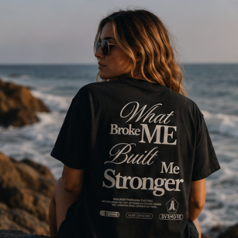 A woman sitting on a rock by the ocean, viewed from behind, wearing a black graphic T-shirt with the text 'What Broke ME Built Me Stronger' in white on the back.