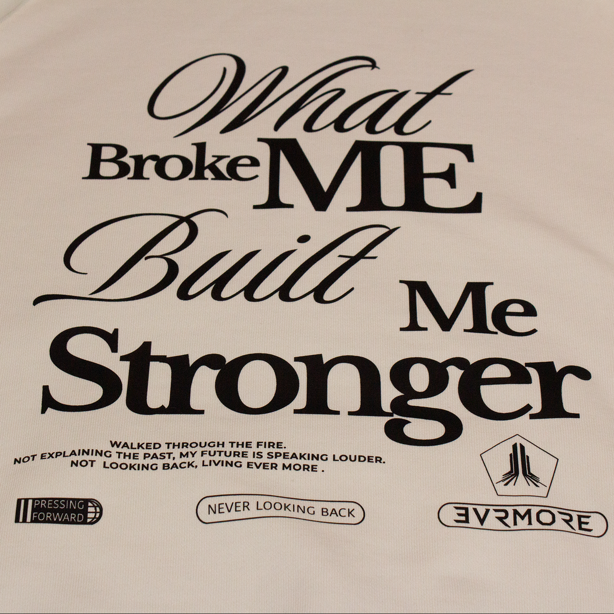 A close-up of a cream-colored t-shirt featuring a black graphic print that reads 'What Broke ME Built Me Stronger' with smaller text and logos below, including 'EVERMORE'.