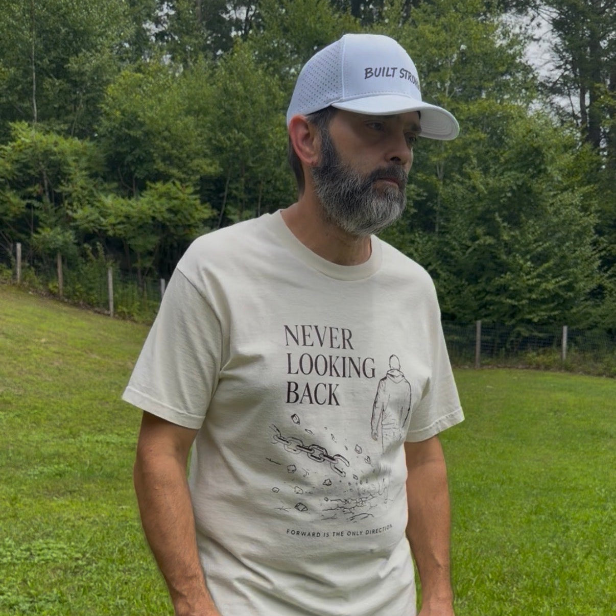 A man wearing a cream-colored graphic t-shirt with the text 'NEVER LOOKING BACK' and a white baseball cap stands in a grassy outdoor field with trees in the background.