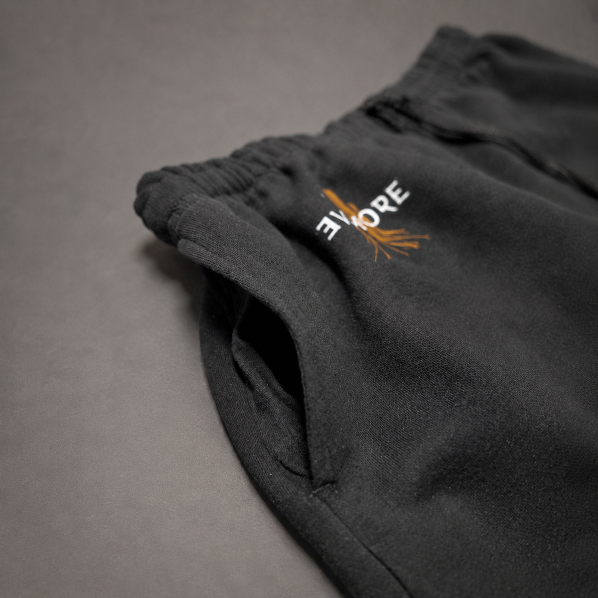 Close-up of dark charcoal gray sweatpants with an elastic waistband, featuring a white and orange embroidered logo on the thigh, set against a neutral gray background.