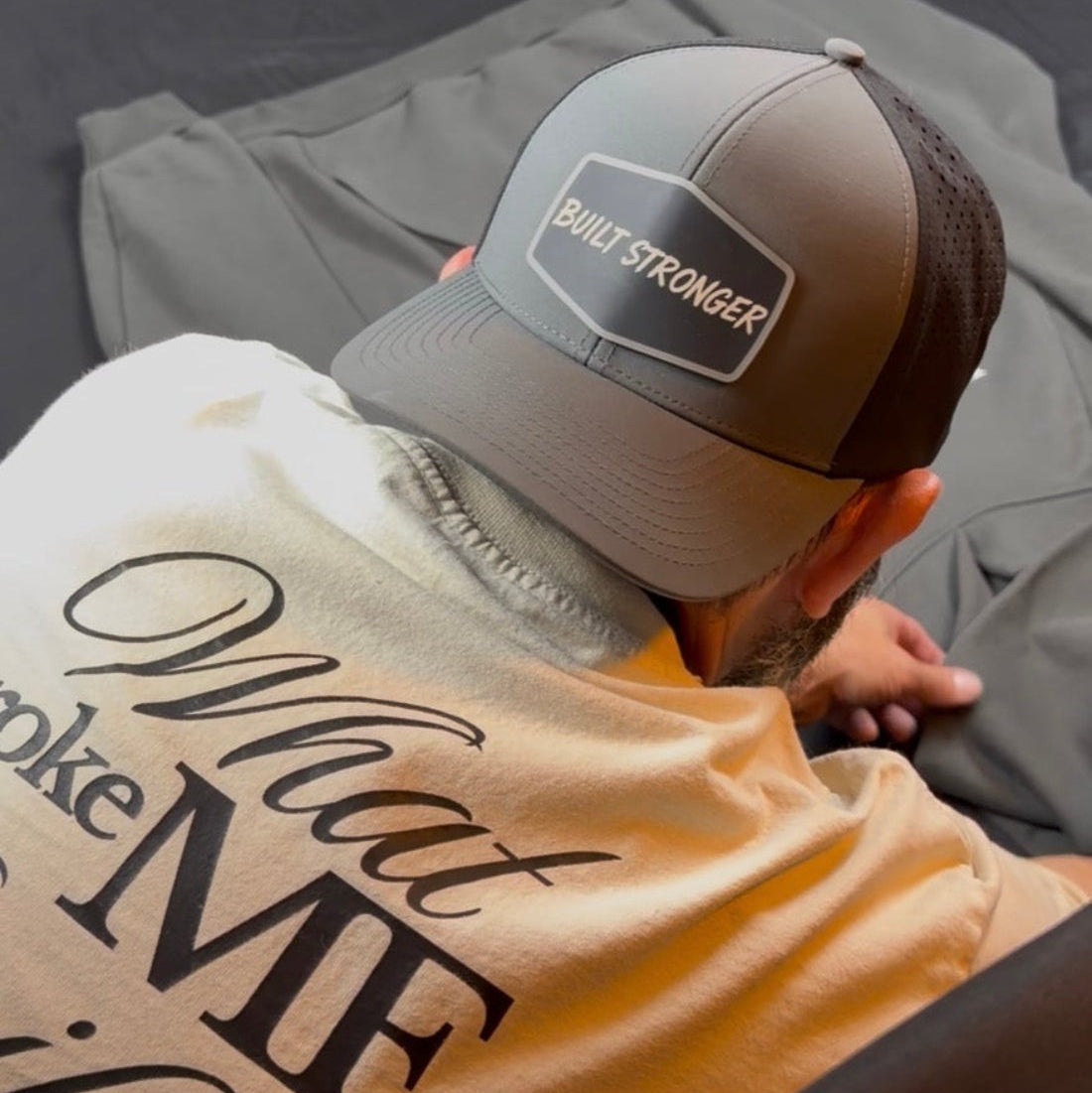A man wearing a gray and black trucker hat with a 'Built Stronger' patch and a beige graphic t-shirt, seen from behind as he sits on a bed with a gray sweatshirt.