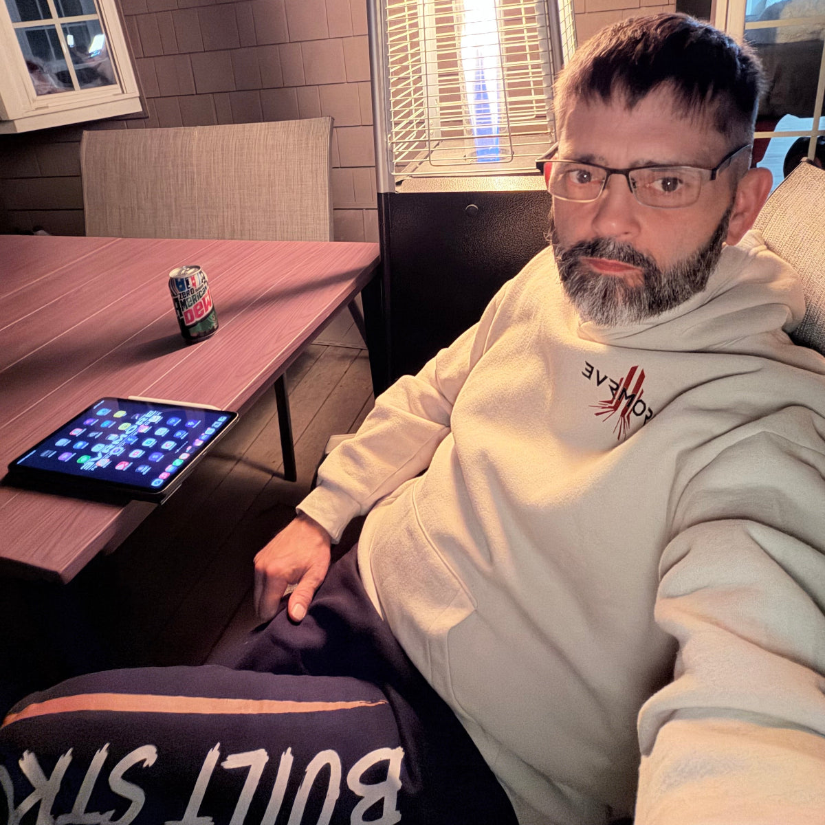 A man wearing a light beige hoodie with a red graphic logo sits on a couch in a dimly lit room, looking at a tablet on a wooden table next to him.